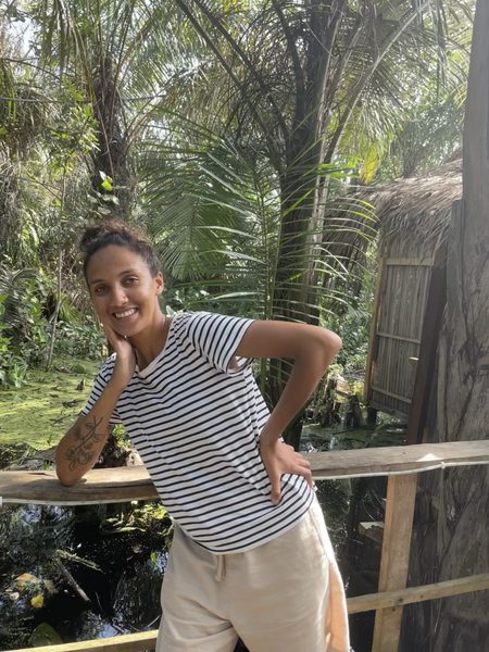 Alicia Telle stands leaning against a wooden railing, green tree fronds in the background