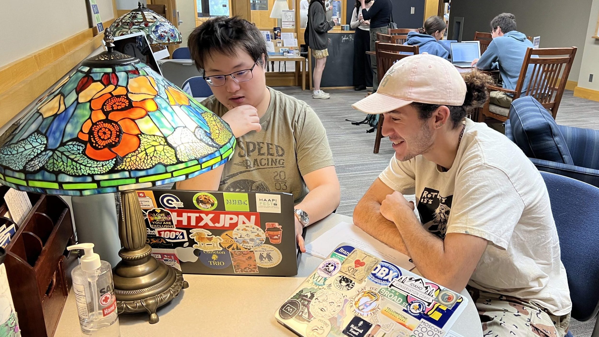 Two students sit side by side at a round table with a computer in front of them.