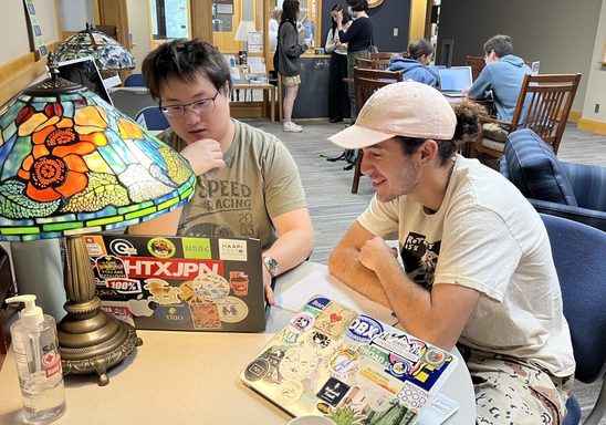 Two students sit side by side at a round table with a computer in front of them.