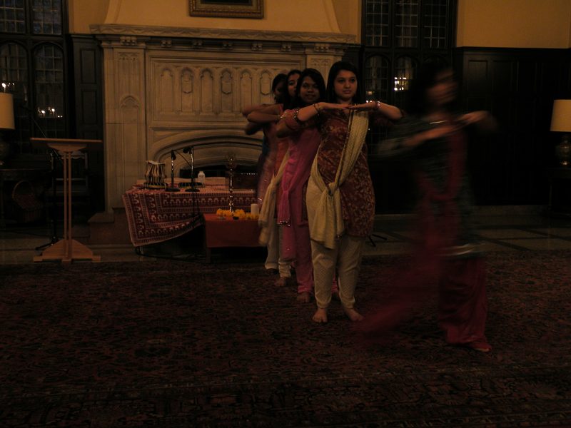 Four dancers in a line with hands raised just below their chin, one dancer in front in a blur from dancing