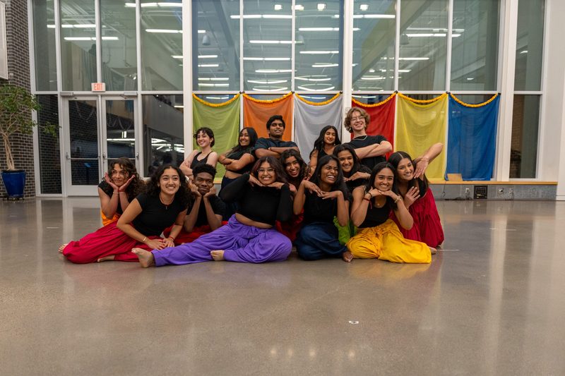 14 student dancers posing posing for a group photo during Diwali 2024, they are wearing bright colored pants with matching black tops