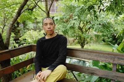 A man in a black shirt with his glasses on his head, sitting on a bench in front of green leaves