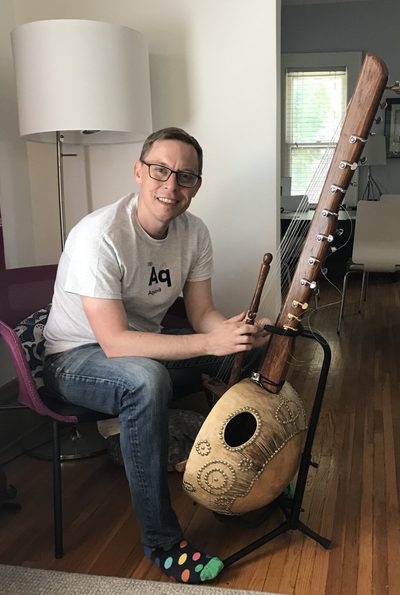 A person sitting on a chair in front of a lamp, with a stringed instruments promped up in front of them.