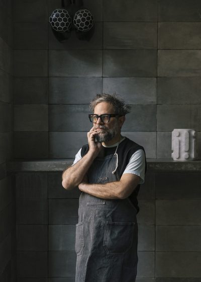Portrait of Pedro Reyes. He is a bearded man with glasses, wearing a denim apron over a shirt and vest. He stands with his arms crossed, one hand touching his chin in thought. Behind him is a wall of large gray square tiles, a concrete shelf with a small sculpture, and two hanging black and white spherical lights.