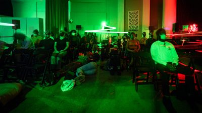 An audience wearing eye masks sits or lies on the floor in an immersive performance space bathed in vibrant green light.