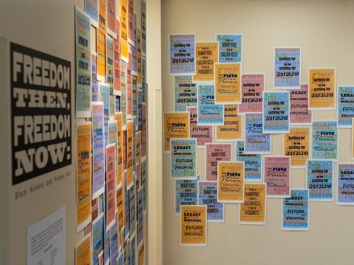 Colorful protest posters overlap and cover two walls. The sign in the foreground reads: Freedom then! Freedom now!