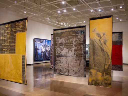 Large yellow, brown, red, and blue prints with photographic and text elements suspended from the ceiling in the Perlman Teaching Museum