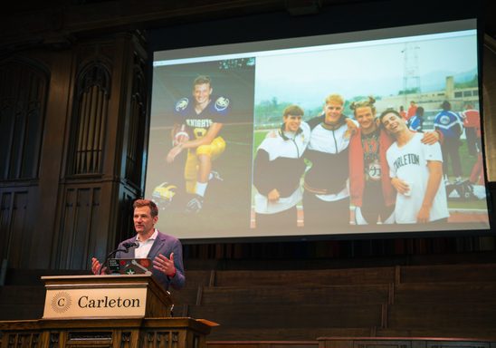 Steve Grove giving his Convo address on Chapel stage. Powerpoint shows two photos of him on the football team while at Carleton.