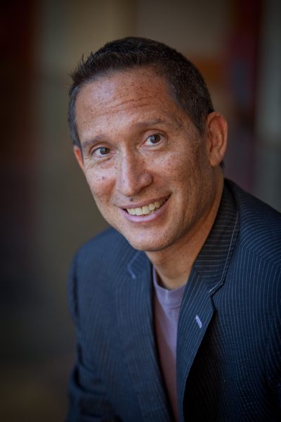 headshot of man with close cropped hair wearing a navy suit jacket over a grey t-shir