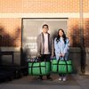 Two student holding green cooler bags filled with food in front of food shelf drop-off door
