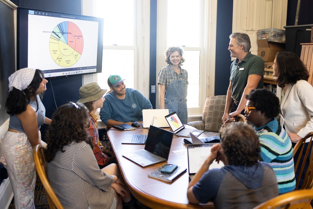 Group of students and educators gathered around a table, laughing. Presentation on a screen.