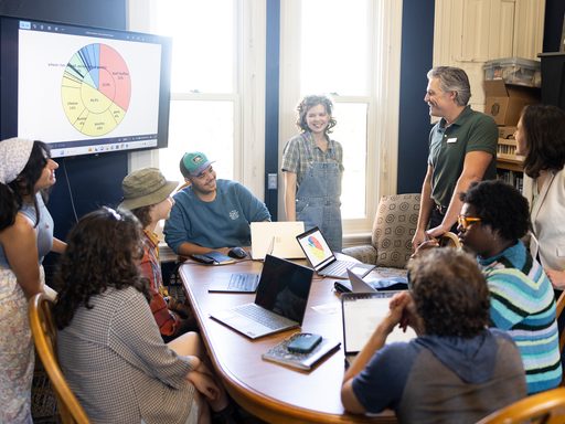 Group of students and educators gathered around a table, laughing. Presentation on a screen.