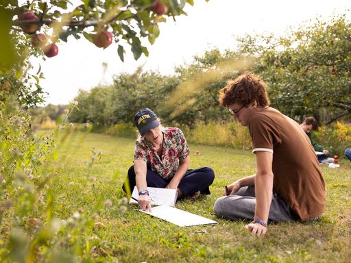 Carleton College professor Eleanor Jensen pointing to a student's sketchbook and giving instructions, while both sit in an apple orchard