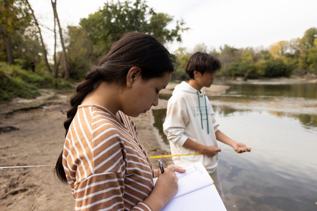 Two Carleton students measuring river water flow, one recording notes in a notebook