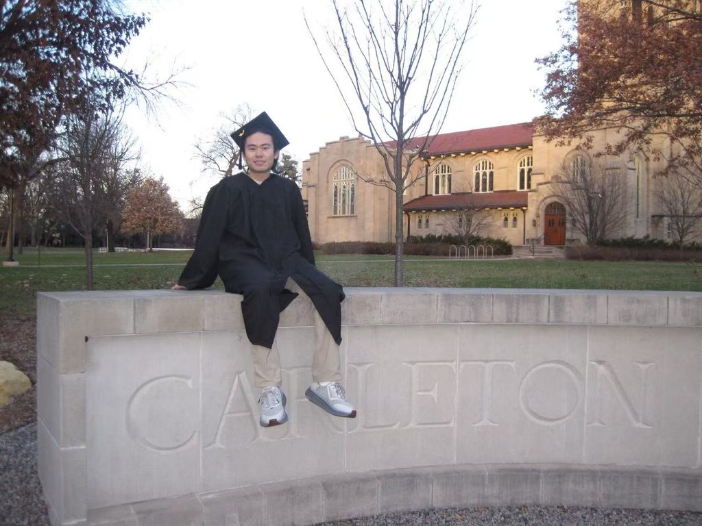james zhang in a graduation cap and gown seated on the stone "carleton" sign