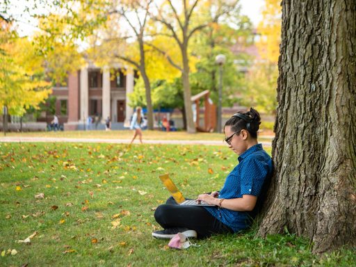 A young person sits outdoors beneath a tree typing on a laptop