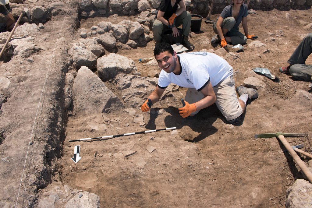 a kneeling student gives a "thumbs up" sign beside some pottery shards and digging tools