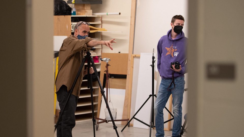 Male professor stands behind a camera with a male student nearby.