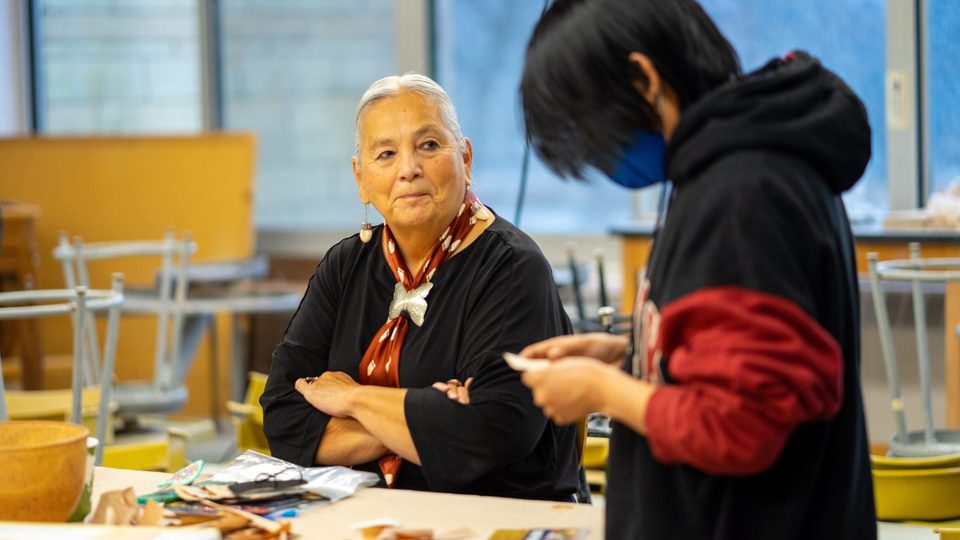 Dr. Denise Lajimodiere, 2022 Elder-in-Residence, sits at a table during her workshop on birch bark-biting. A student in the foreground is looking down at their piece of bark.