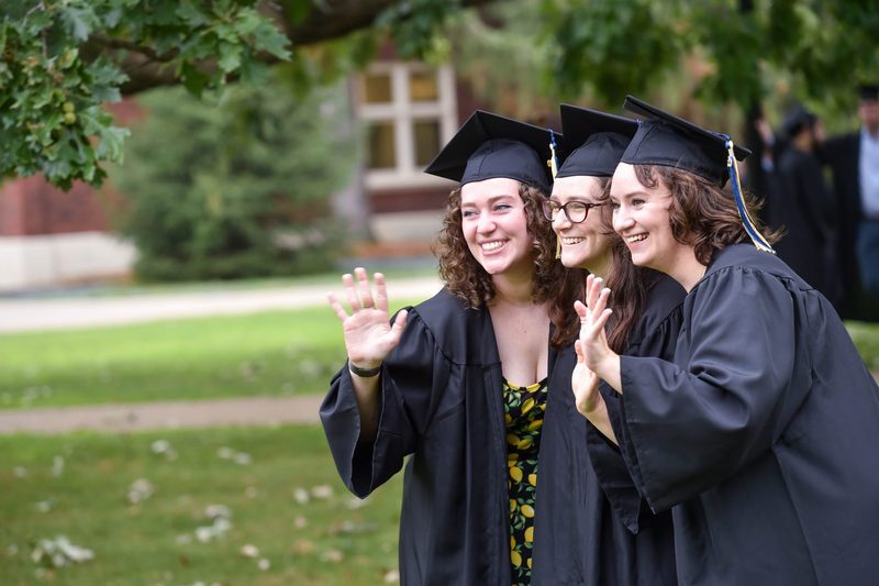 Erica Helgerud '20, Katie Paasche '20 and Kyra Wilson '20, all in graduation caps and gowns, wave at a different camera.