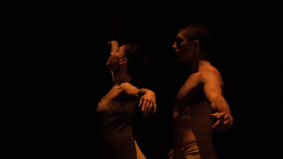 Two ballet dancers perform a duet on a very dark stage, barely lit by warm light.