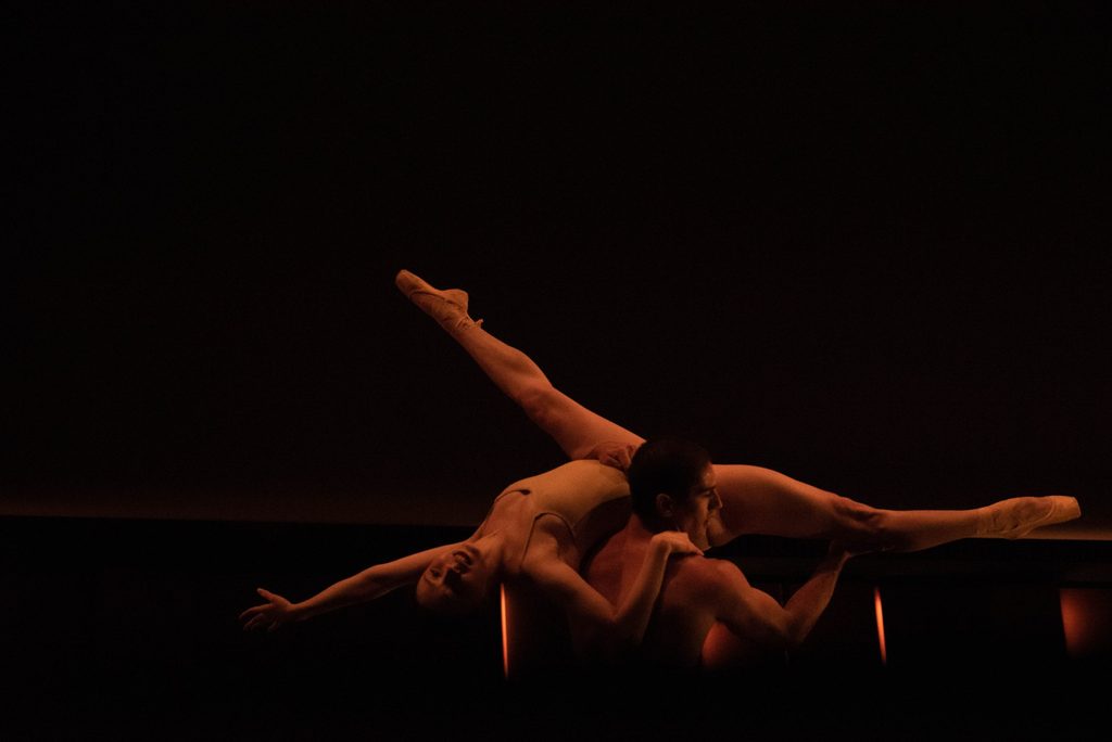 One ballet dancer lifts another upside down as they perform a duet on a very dark stage, barely lit by warm light.