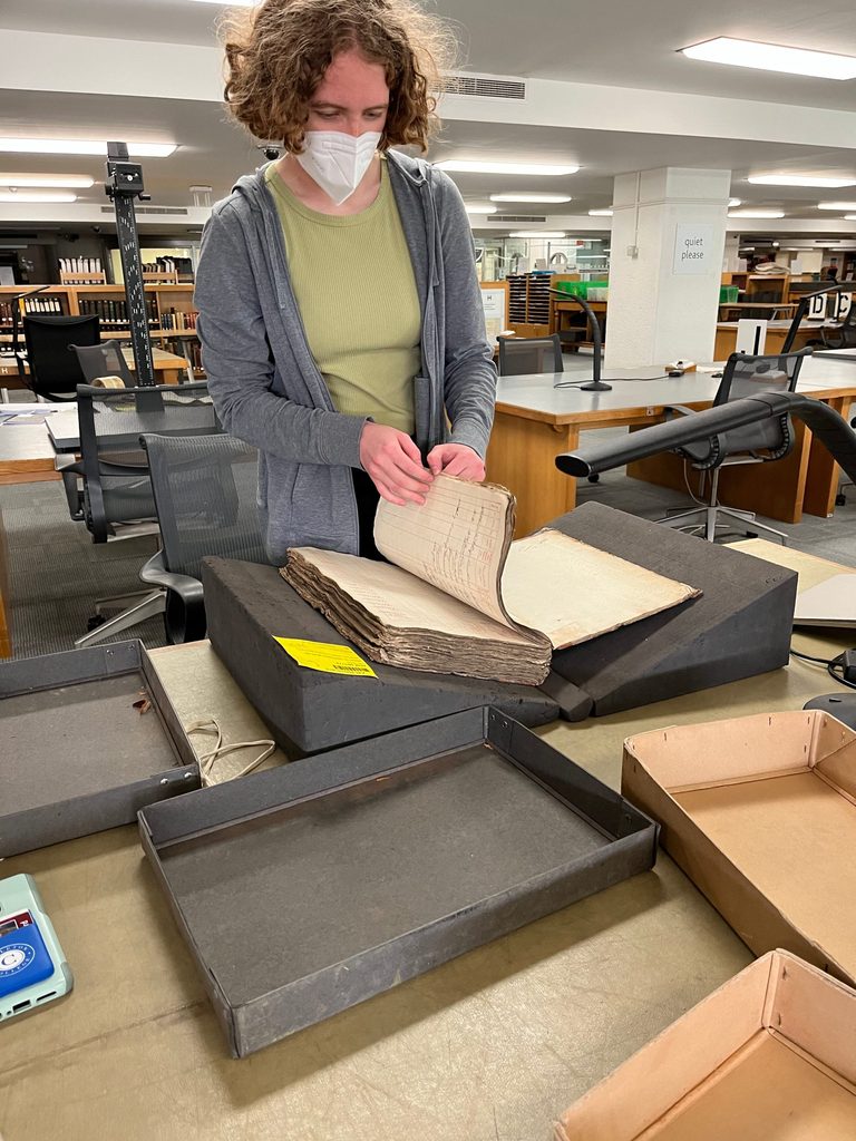 In a library, Raine Bernhard ’23 turns the pages of a very old, thick book.