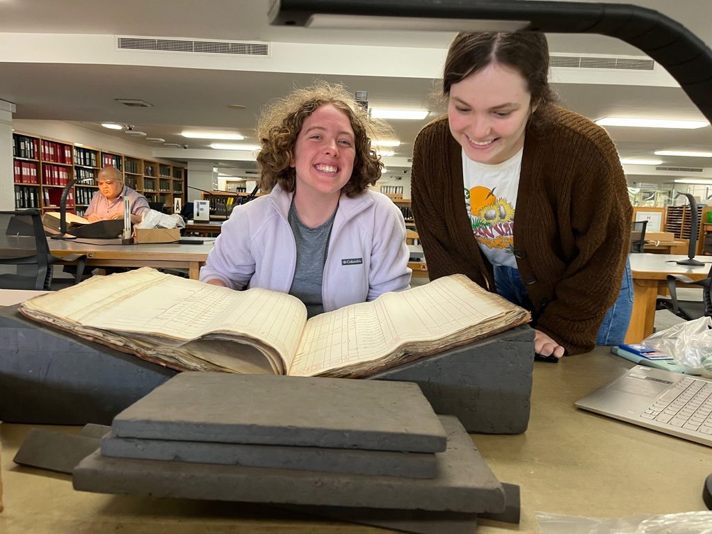 Raine Bernhard ’23 smiles at the camera and Margaret De Fer ’24 smiles down at a large, very old ledger book.