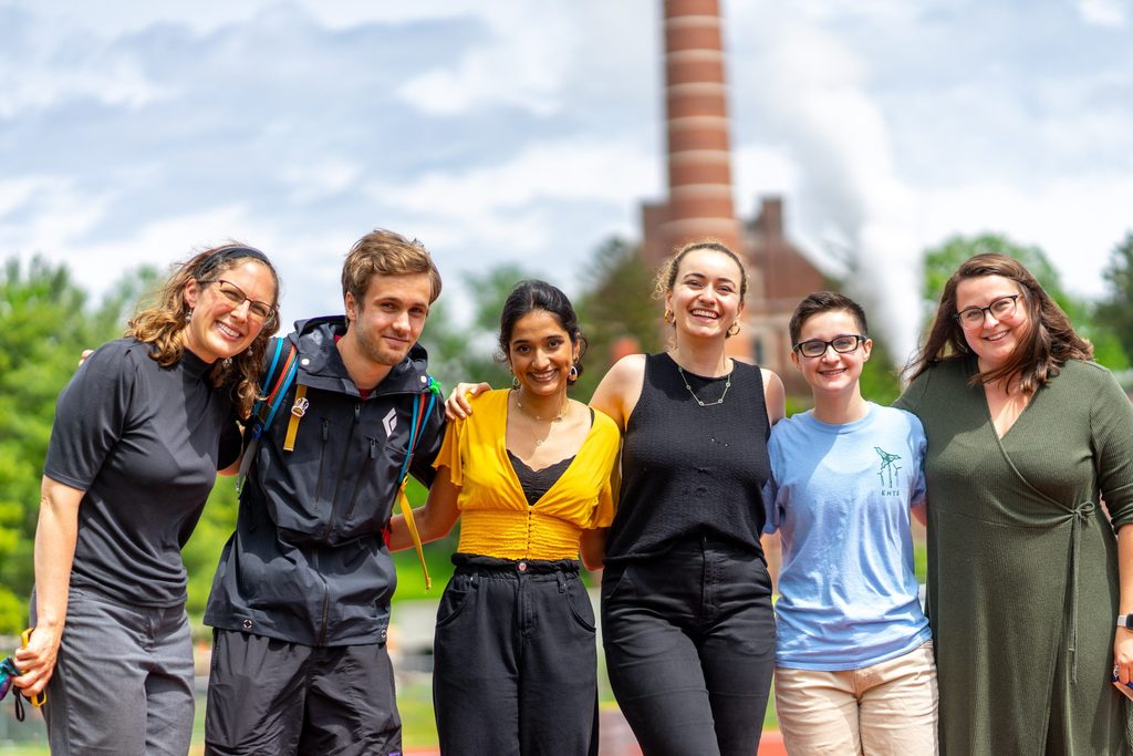 Four students and two staff members pose together in front of the Carleton steam stack.