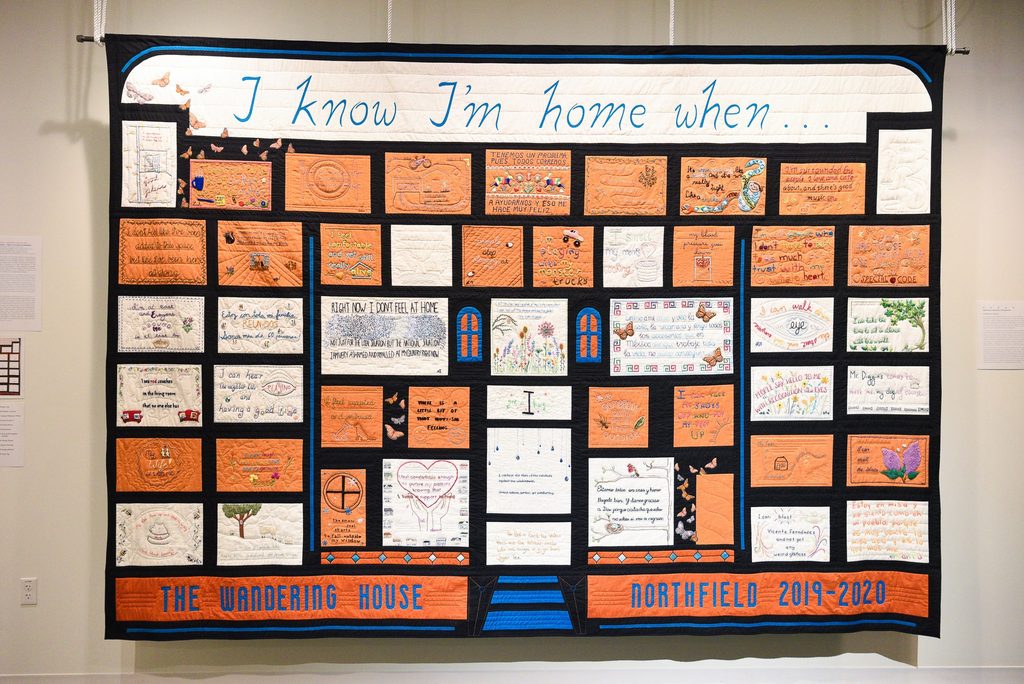 Large quilt hanging on an exhibit wall. Most panels are orange or white, with multicolored embroidery. "I know I'm home when..." is embroidered at the top and "The Wandering House Northfield 2019-2020" at the bottom.