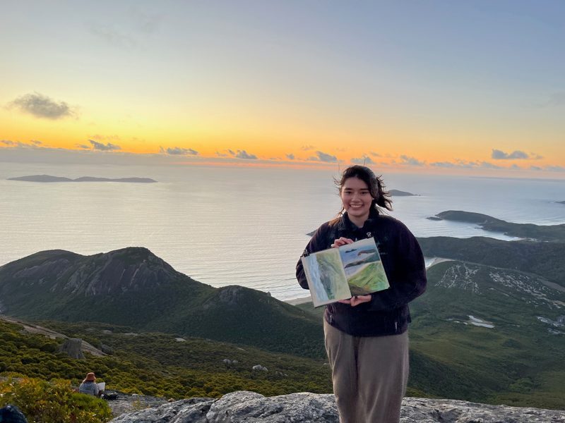 student poses with sketchbook in front of sunset