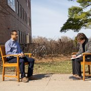 A professor and a student sit opposite each other on two benches, outside in the sun.