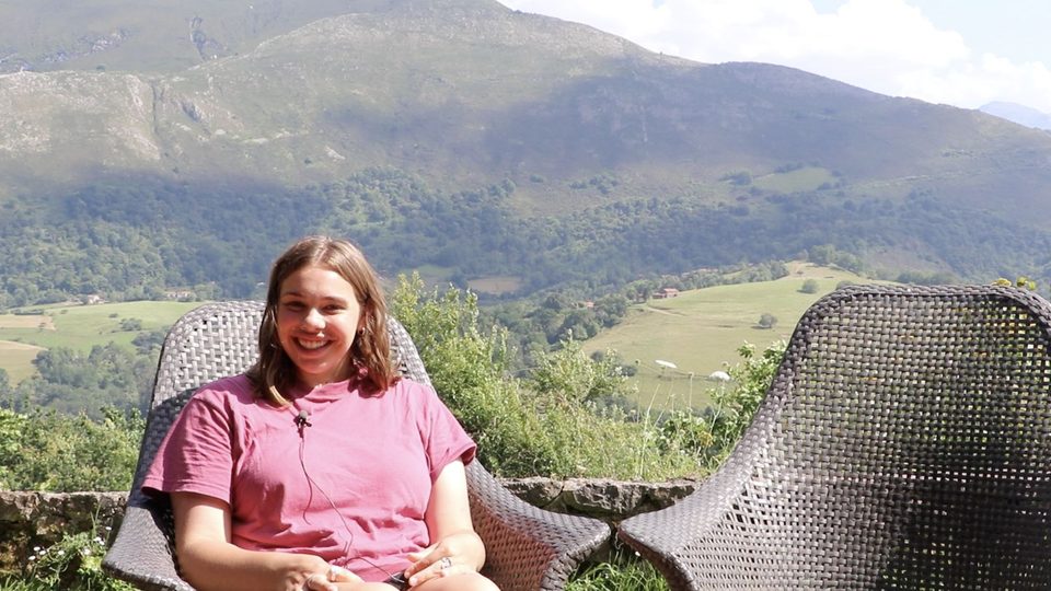 A student smiles as she sits in a chair, with a green valley and mountains behind her.