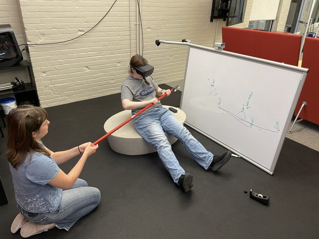 Two students test a virtual reality rowing game with a broom handle