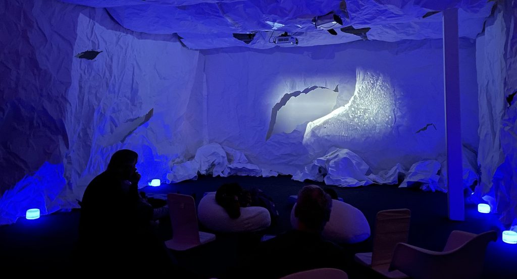 People visiting the installation sit and watch. The room is filled with blue light and a picture of cracking ice is on the wall