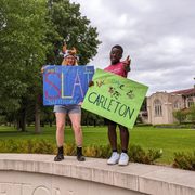 Two Carleton students stand on the Carleton sign, one holding a 