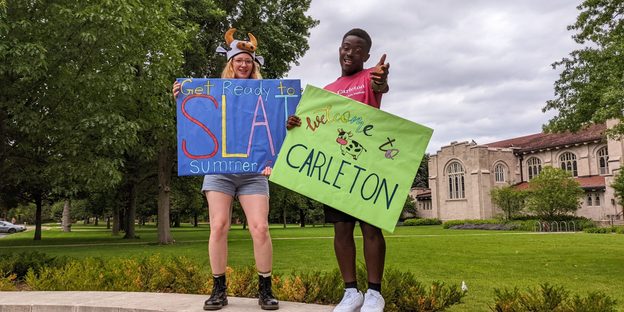 Two Carleton students stand on the Carleton sign, one holding a 
