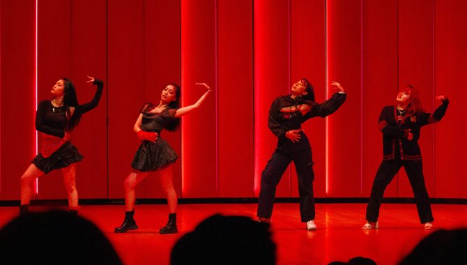 Four students in coordinated black outfits mid-dance onstage in Kracum Performance Hall, with deep red lights behind them.