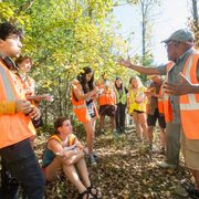 Professor Bereket Haileab gestures widely as he speaks to a group of students in a forest. All wear neon orange or yellow safety vests.