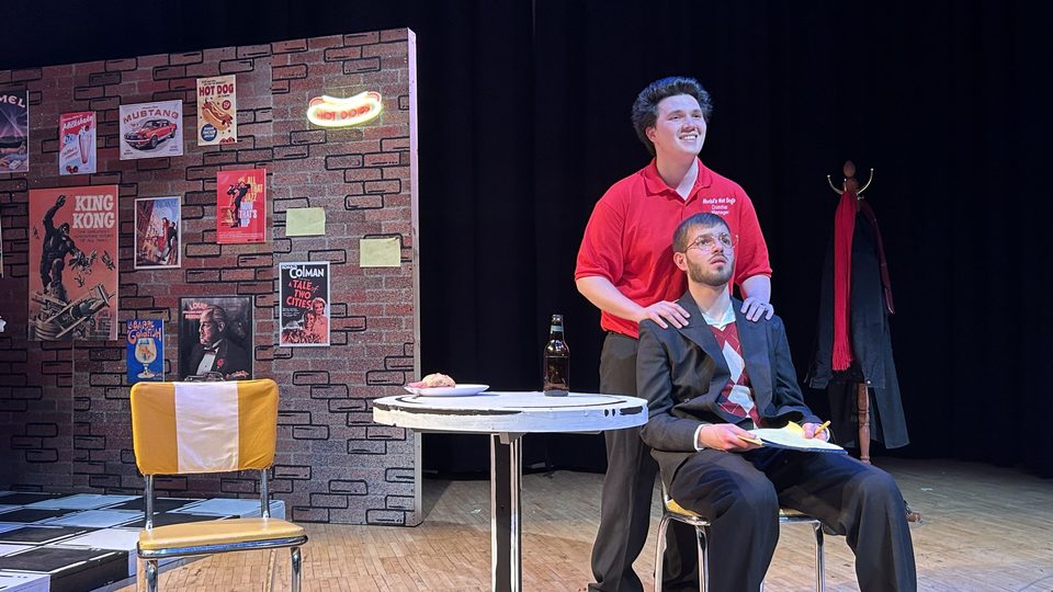 Preves as Louis sits in a chair, looking out at the audience while Le as Cruncher speaks and rests his hands on Louis' shoulders.