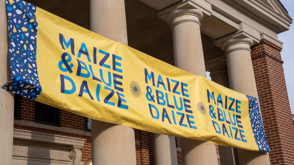 Close-up on the Maize & Blue Daize banner attached to the columns of Sayles-Hill Campus Center.