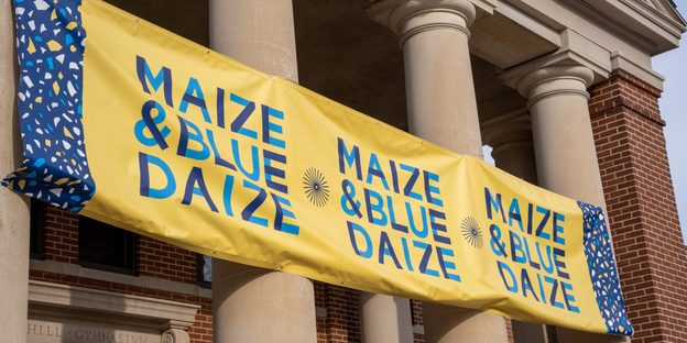 Close-up on the Maize & Blue Daize banner attached to the columns of Sayles-Hill Campus Center.