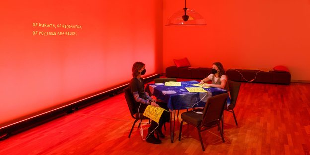 Two people sitting at a table in a room lit with red light, heating pads and cords are strewn about chairs and benches. On the wall is projected a transcription of an audio installation of Finnegan Shannon and Ezra Benus that addresses the culture around heating pads.