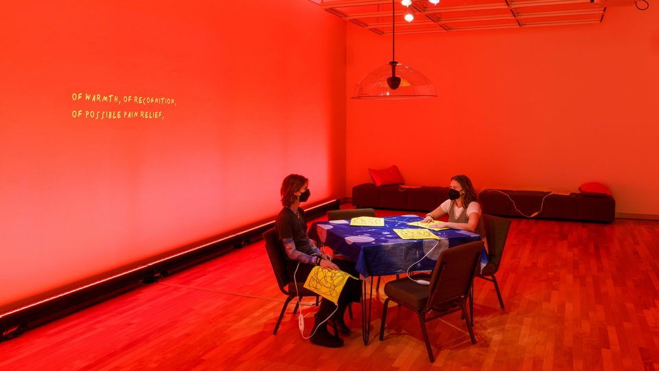 Two people sitting at a table in a room lit with red light, heating pads and cords are strewn about chairs and benches. On the wall is projected a transcription of an audio installation of Finnegan Shannon and Ezra Benus that addresses the culture around heating pads.