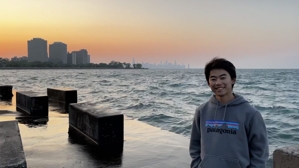 Kai Oishi standing outside by a body of water during sunset, with a city skyline in the background.