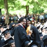 Two students in graduation gowns stand and hug in the midst of other, sitting students in graduation gowns.