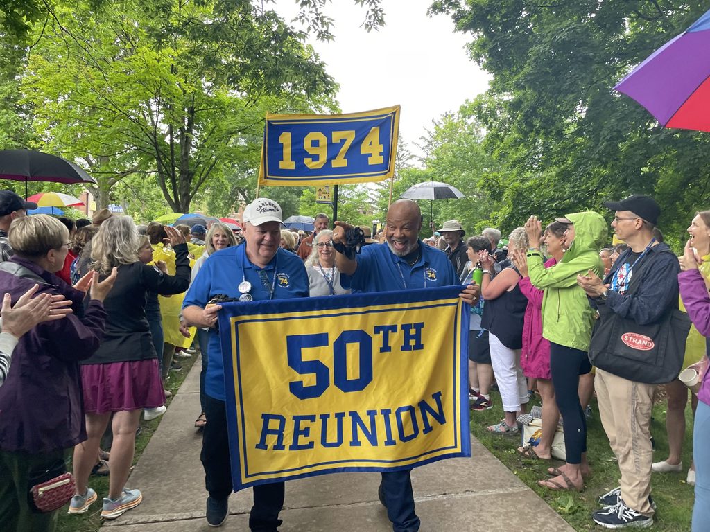 Two male members of the Carleton Class of 1974 carry a banner that says, "50th Reunion."