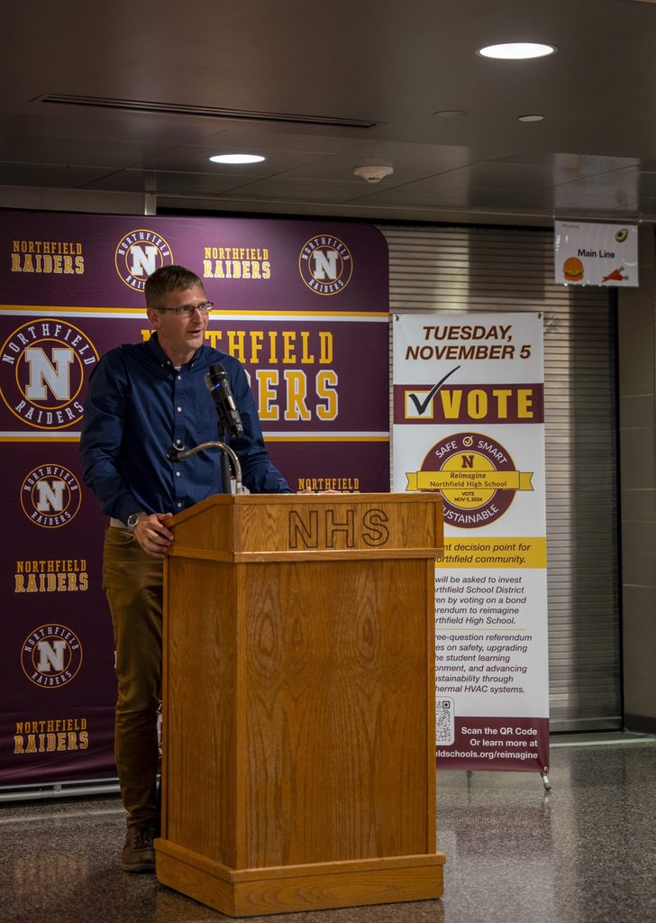 Carleton professor Matt Whited speaks at a podium in front of a Northfield Raiders banner.