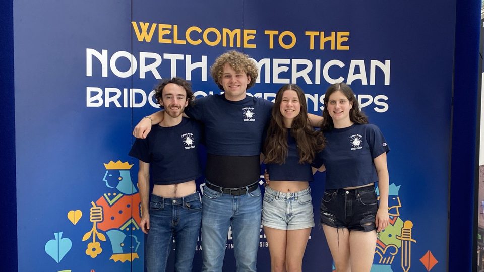 Four Carleton students in matching T-shirts pose in front of a sign that says, 