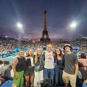 Six people pose in the crowd at an Olympic volleyball game, with the Eiffel Tower in the background, right after the sun sets.
